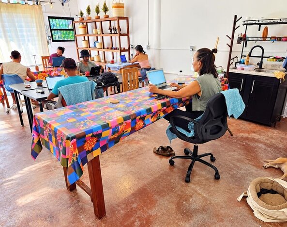 Spacious coworking area with several people working on laptops at colorful communal tables, assorted chairs, wooden display shelving, kitchenette, and a dog resting on the floor.
