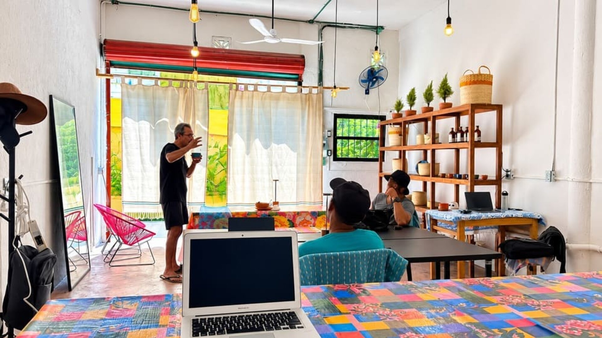 Colorful coworking area with people working and talking around communal tables, laptops, bright woven chairs, hanging lights, wooden shelving, and large windows.