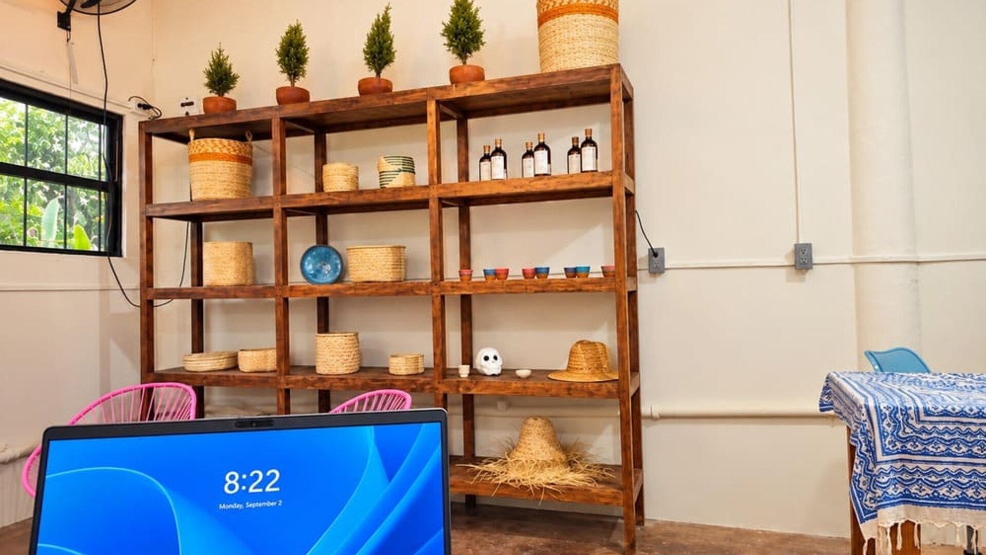 Bright coworking space with wooden shelving displaying woven baskets, small potted plants, ceramics, and bottles, with a computer workstation in the foreground.
