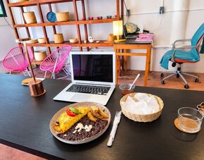 Open laptop beside a plated breakfast of omelet, black beans, potatoes, and cheese on a spacious coworking table with colorful seating and decorative shelving.