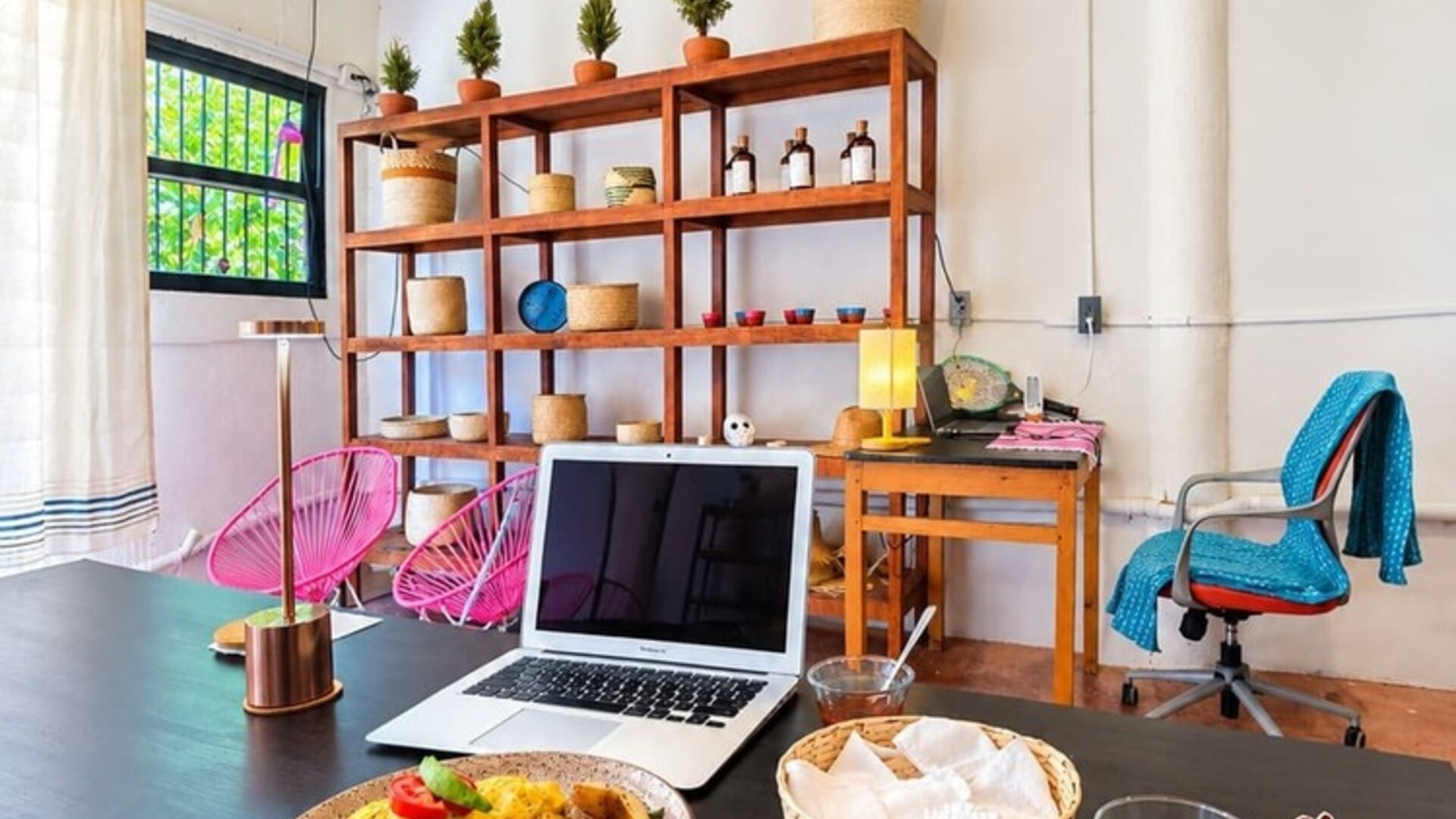 Bright coworking area with an open laptop and breakfast on a large black table, surrounded by pink chairs, wooden shelving, woven décor, plants, and warm pendant lighting.