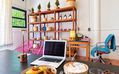 Bright coworking area with an open laptop and breakfast on a large black table, surrounded by pink chairs, wooden shelving, woven décor, plants, and warm pendant lighting.