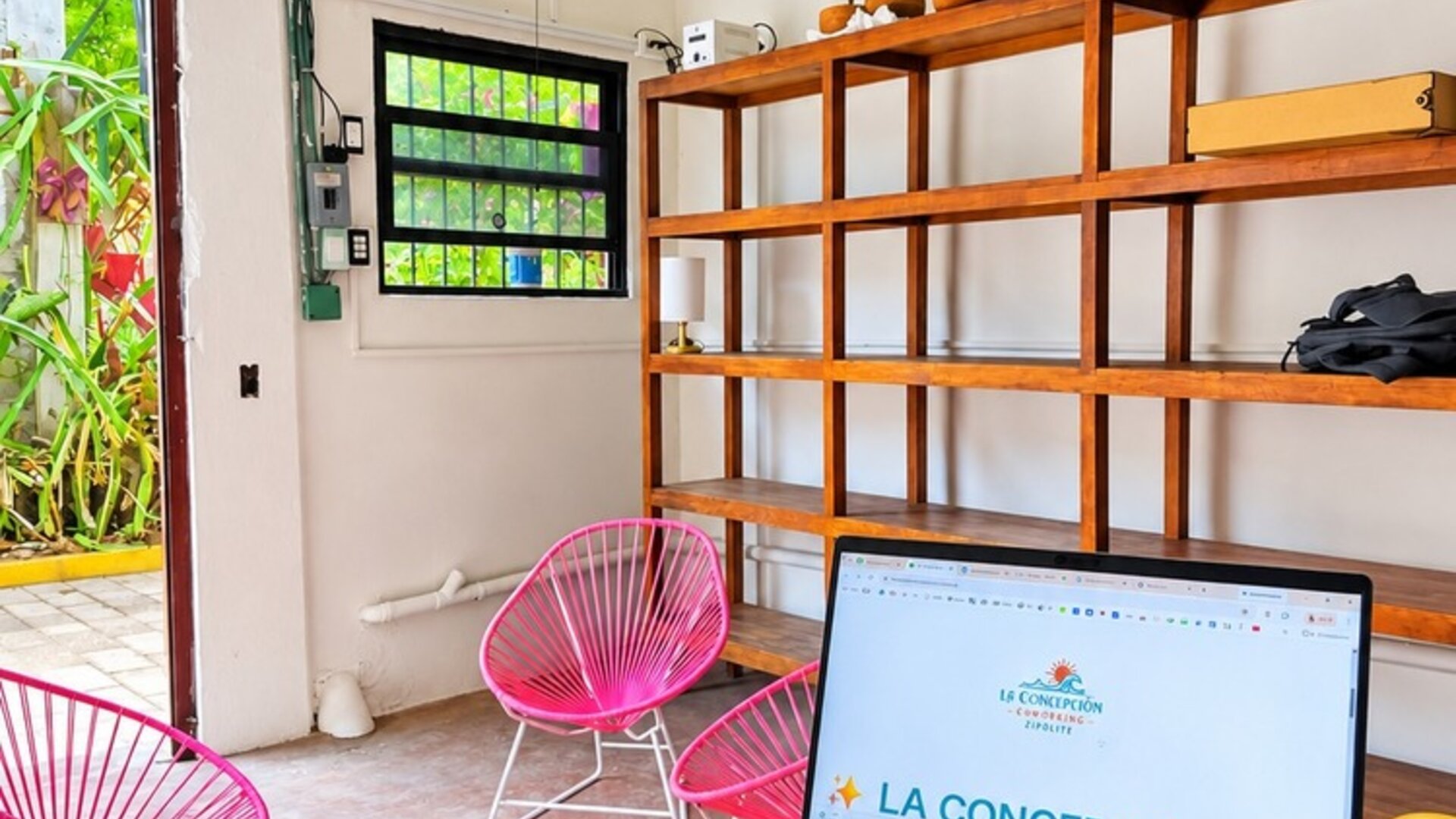 Laptop on a black table in a bright coworking space with pink woven chairs, wooden shelving, potted plants, large windows, and an open doorway to greenery.