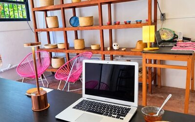 Breakfast plate with an omelet, black beans, potatoes, and fresh garnishes beside an open laptop in a bright coworking space with pink chairs and wooden shelving.