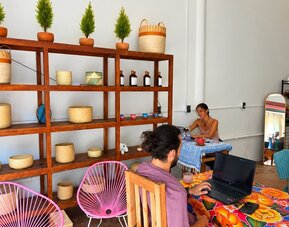 Relaxed coworking space with people working on laptops at colorful tables, pink woven chairs, decorative wooden shelving, potted plants, and warm pendant lighting.
