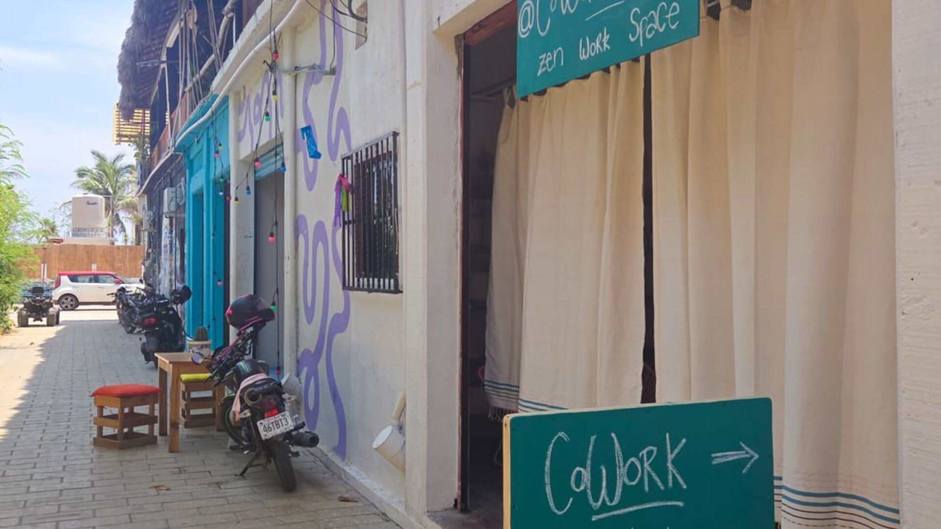 Street view of a relaxed coworking space entrance with turquoise signage, white curtains, parked motorcycles, and neighboring buildings along a sunny pedestrian lane.