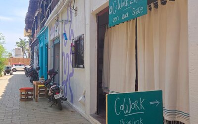 Street view of a relaxed coworking space entrance with turquoise signage, white curtains, parked motorcycles, and neighboring buildings along a sunny pedestrian lane.