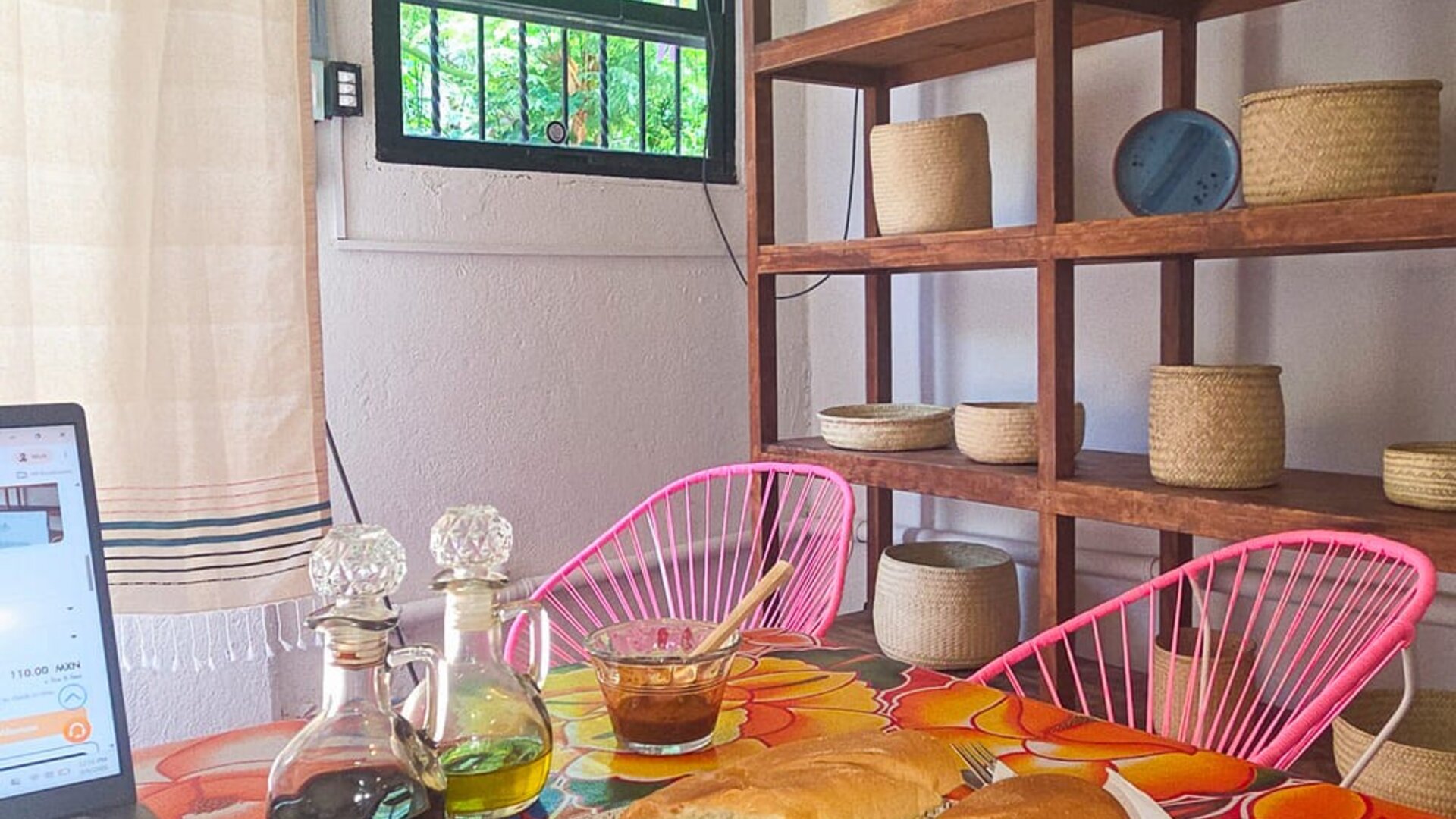 Sandwich and green salad served beside a laptop on a vibrant floral table, with condiment bottles, pink woven chairs, wooden shelving, and natural light.