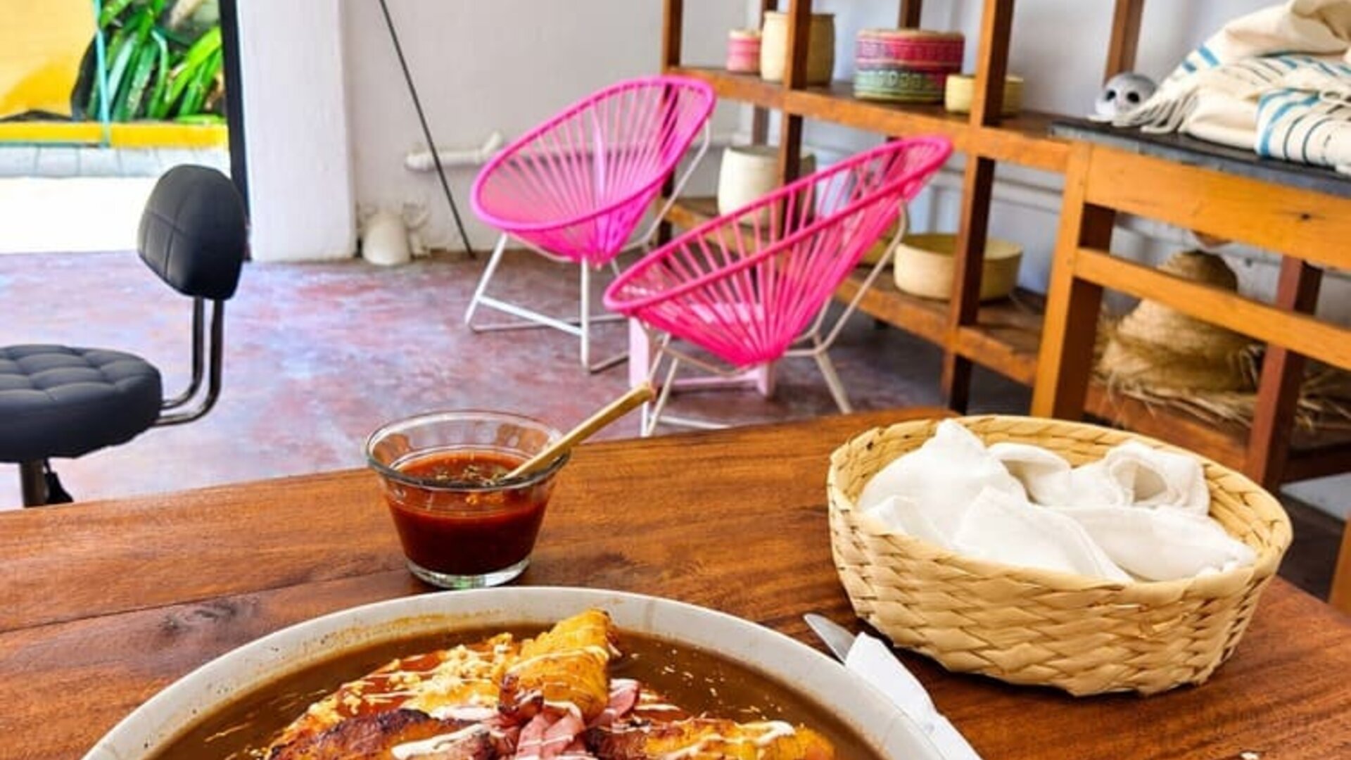 Plate of Mexican-style food with fried plantains, sauce, cheese and toppings served on a table with salsa and napkins, surrounded by wooden shelving, woven décor, bright pink chairs, and a window overlooking tropical greenery.