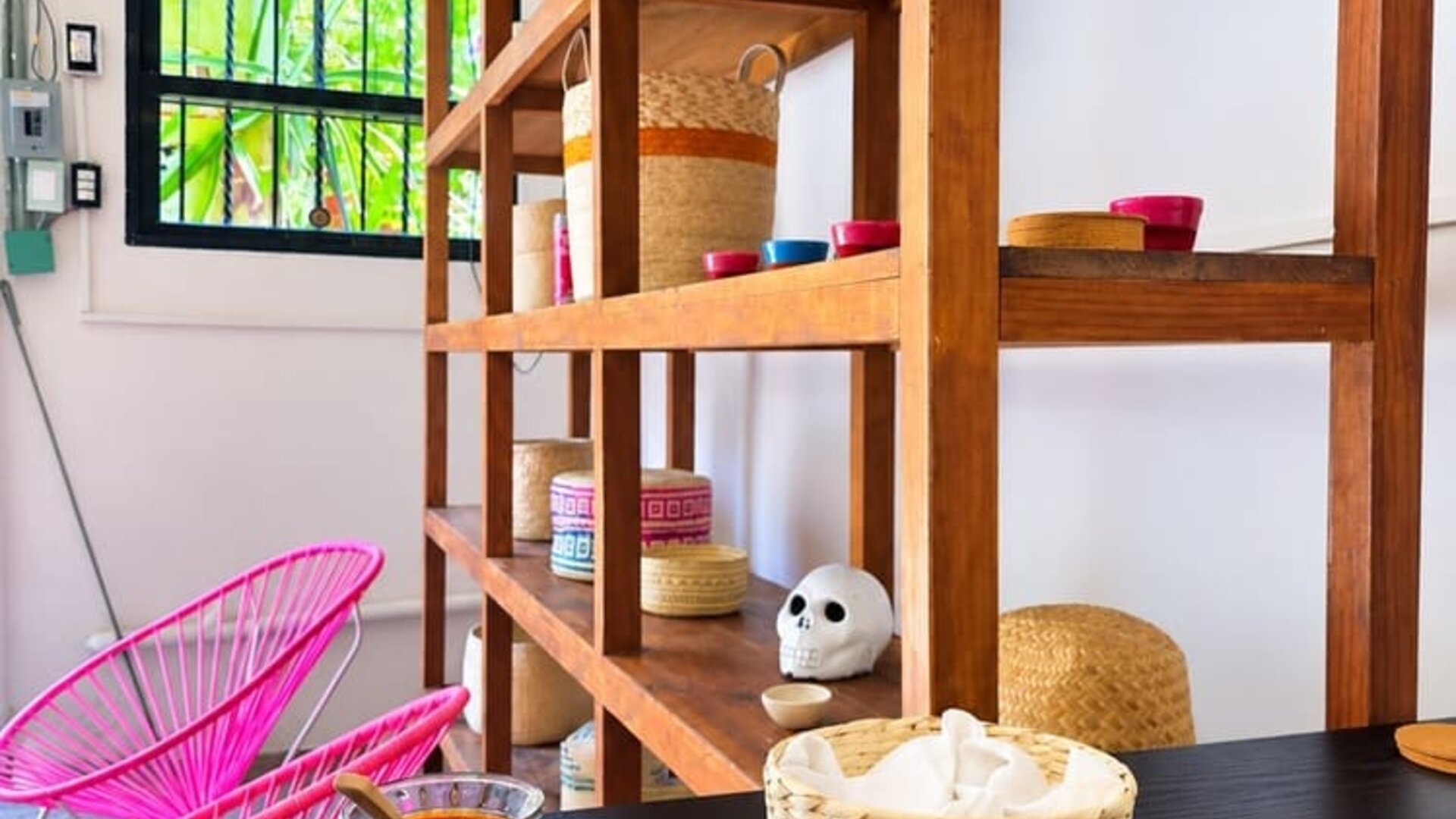 Colorful coworking café interior with a large plate of Mexican-style food and fried plantains on a wooden table, salsa and napkins, bright pink woven chairs, wooden shelving, and tropical greenery visible outside.