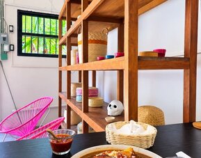 Colorful coworking café interior with a large plate of Mexican-style food and fried plantains on a wooden table, salsa and napkins, bright pink woven chairs, wooden shelving, and tropical greenery visible outside.