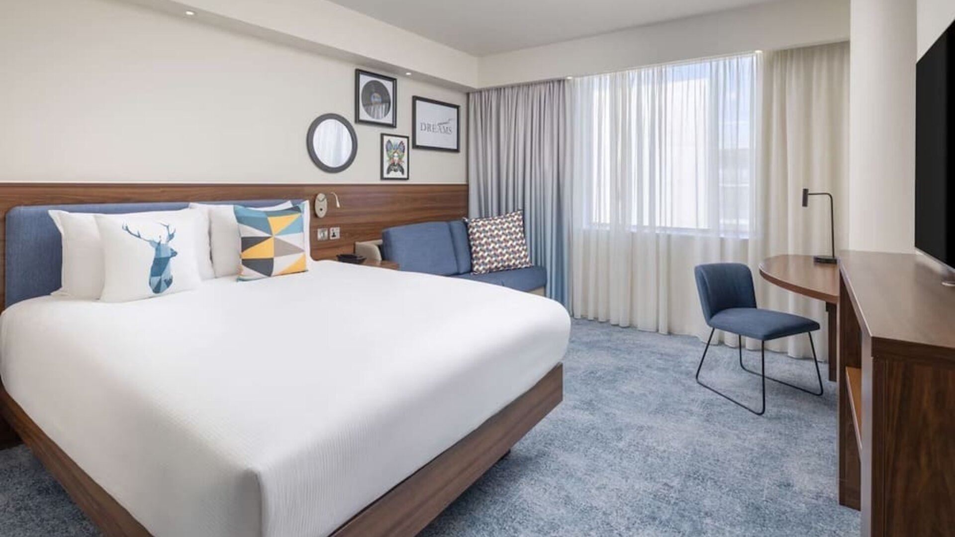 Bright contemporary king room with a white bed, blue upholstered headboard, colorful pillows, sofa, work desk, chair, large windows and wall-mounted TV.