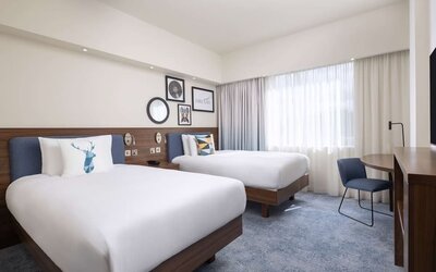 Spacious hotel twin room with two white beds, blue upholstered headboards, decorative pillows, large curtained windows, work desk, chair and wooden TV console.