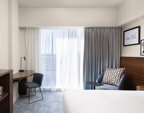 Bright modern hotel room with a large bed, floor-to-ceiling window and curtains, blue armchair, wooden work desk, chair, wall-mounted TV and contemporary artwork.