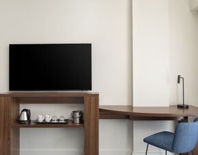 Hotel room workspace with a large flat-screen TV, curved wooden desk, blue chair, desk lamp and hospitality station with kettle, cups, glasses and ice bucket.