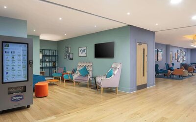Colorful contemporary hotel lobby with teal and patterned lounge chairs, small tables, pale wood flooring, pastel green and blue walls and multiple seating areas.