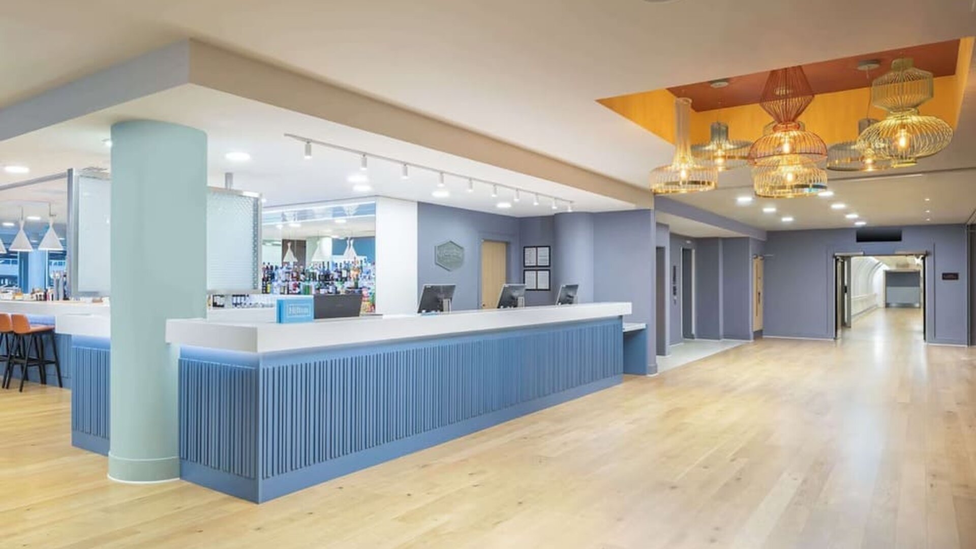 Spacious modern hotel reception area with a long blue front desk, light wood flooring, pastel blue walls, decorative pendant lights and an adjoining bar.