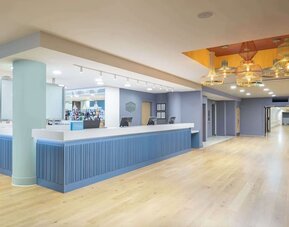 Spacious modern hotel reception area with a long blue front desk, light wood flooring, pastel blue walls, decorative pendant lights and an adjoining bar.