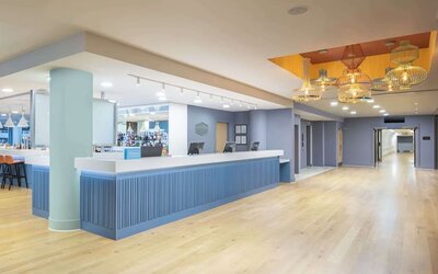 Spacious modern hotel reception area with a long blue front desk, light wood flooring, pastel blue walls, decorative pendant lights and an adjoining bar.