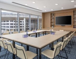 Bright hotel meeting room with U-shaped conference tables, upholstered chairs, large wall-mounted TV, wooden shelving and floor-to-ceiling windows overlooking nearby buildings.