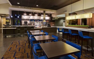 Contemporary hotel café and bar area with blue chairs, dark dining tables, counter seating, pendant lights and a service counter with drinks and snacks.