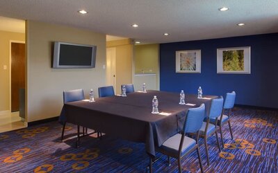 Hotel meeting room with rectangular conference tables, blue chairs, bottled water, wall-mounted flat-screen TV, framed artwork and patterned blue-and-orange carpet.