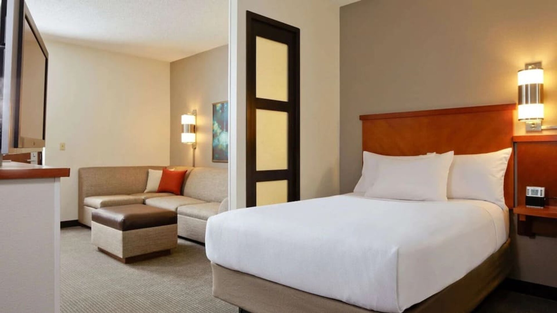 Spacious hotel day room with a king bed, separate sitting area with a sofa and ottoman, work desk, wall-mounted TV, large window, and warm contemporary décor.