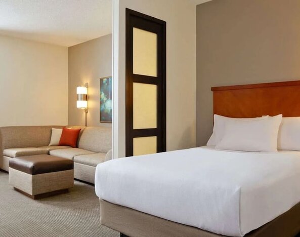 Spacious hotel day room with a king bed, separate sitting area with a sofa and ottoman, work desk, wall-mounted TV, large window, and warm contemporary décor.