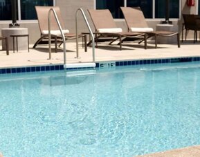 Hotel outdoor swimming pool with clear blue water, metal entry ladder, spacious pool deck, cushioned lounge chairs, side tables, and additional outdoor seating.