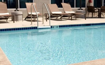 Hotel outdoor swimming pool with clear blue water, metal entry ladder, spacious pool deck, cushioned lounge chairs, side tables, and additional outdoor seating.