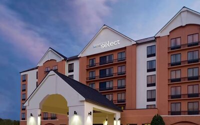 Exterior view of the multi-story Sonesta Select hotel at dusk, featuring a peach and cream façade, illuminated hotel signage, balconies, and a covered entrance.