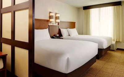 Spacious hotel room with two double beds dressed in white linens, wooden headboards, contemporary wall lights, a large curtained window, neutral carpeting, and a decorative room divider.