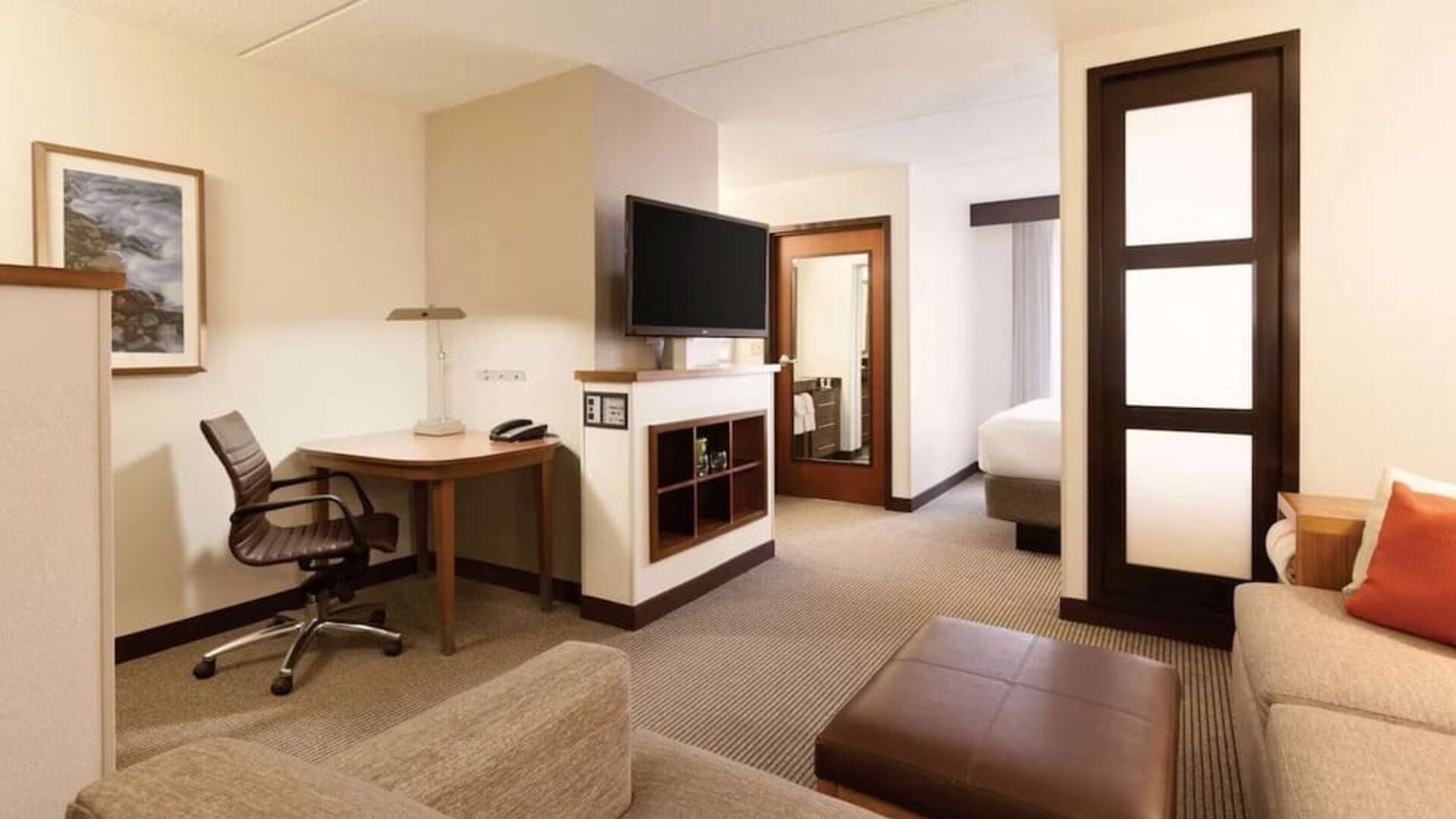 Spacious hotel room featuring a king bed with white linens, a separate sitting area with sectional sofa and ottoman, contemporary wood accents, wall lighting, and a partially visible TV.