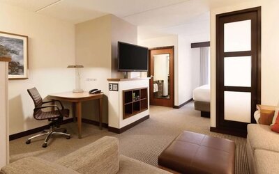 Spacious hotel room featuring a king bed with white linens, a separate sitting area with sectional sofa and ottoman, contemporary wood accents, wall lighting, and a partially visible TV.
