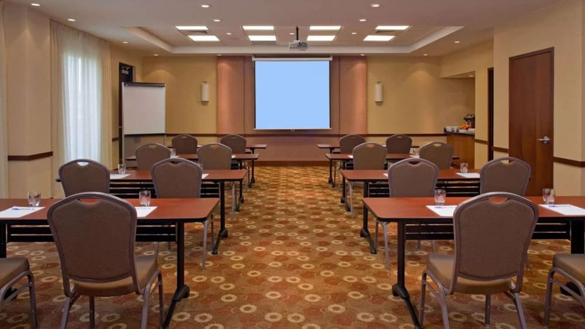 Hotel meeting and conference room arranged classroom-style with tables and chairs, a large projection screen, flip chart, ceiling-mounted projector, and refreshment station.