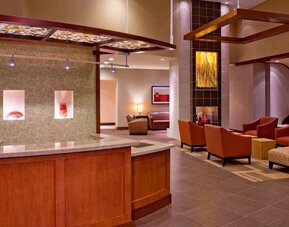 Contemporary hotel reception and lobby area featuring a wood and granite front desk, rust-colored lounge chairs, decorative wall art, modern overhead fixtures, and warm ambient lighting.