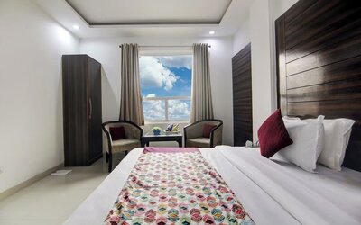 Bright hotel guest room with a neatly made bed featuring white linens, burgundy accent pillows and a colorful patterned runner, plus a wall-mounted TV, wardrobe, two armchairs, small table, ceiling fan and large window.