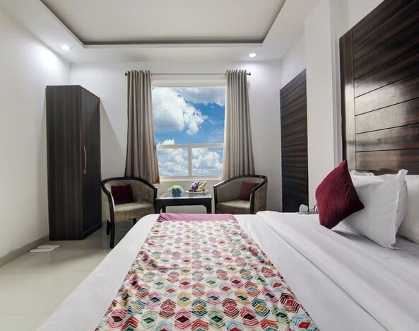 Bright hotel guest room with a neatly made bed featuring white linens, burgundy accent pillows and a colorful patterned runner, plus a wall-mounted TV, wardrobe, two armchairs, small table, ceiling fan and large window.