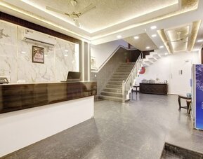 Spacious hotel lobby with a dark wood reception counter, marble-patterned wall, illuminated ceiling, gray tiled staircase with glass railing, seating area and beverage cooler.