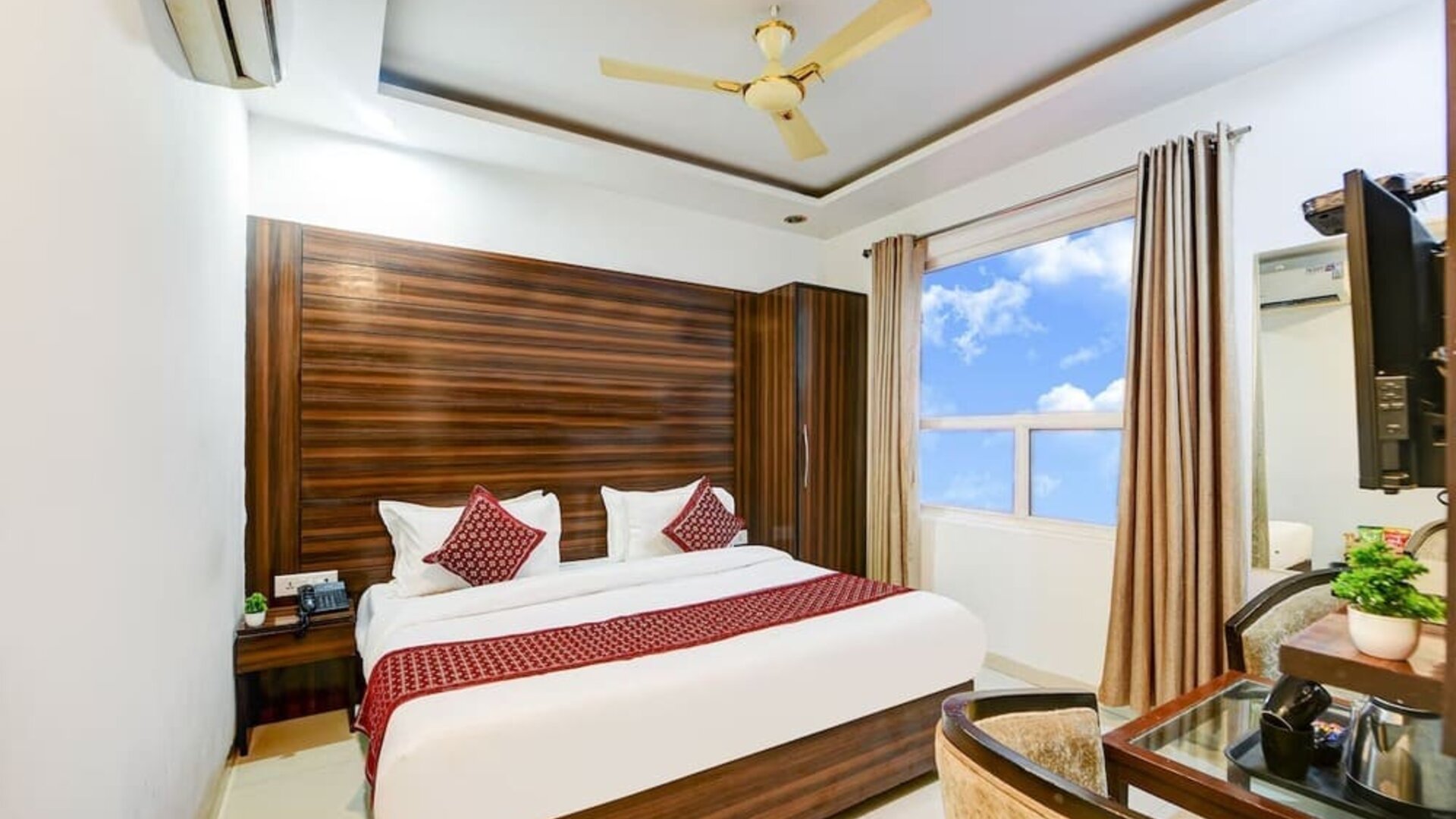 Bright hotel room with a large bed dressed in white linens, burgundy pillows and a colorful patterned runner, complemented by a dark wood headboard, wardrobe, two armchairs, small table and large window with curtains.