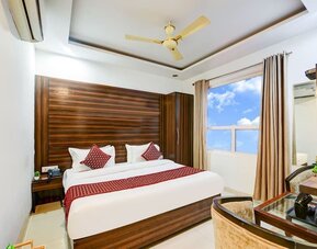 Bright hotel room with a large bed dressed in white linens, burgundy pillows and a colorful patterned runner, complemented by a dark wood headboard, wardrobe, two armchairs, small table and large window with curtains.