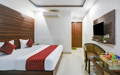 Contemporary hotel room with a large bed featuring white linens and red patterned accents, dark wood wall panels, wall-mounted TV, air conditioning, upholstered seating and a glass table with refreshments.