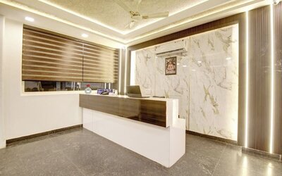 Modern hotel reception desk with dark wood and white finishes, marble-patterned feature wall, vertical accent lighting, large window with striped blinds, ceiling fan and polished gray flooring.
