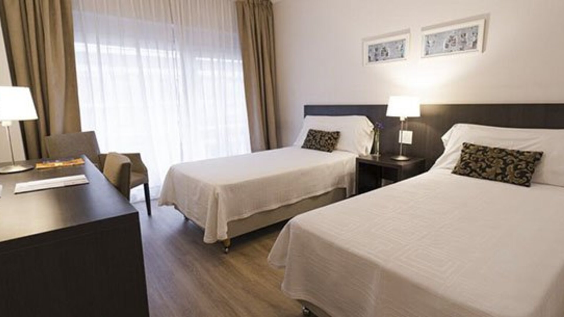 Twin-bed guest room with a desk, television, armchair, bedside table, and large curtained window at El Conquistador Hotel.