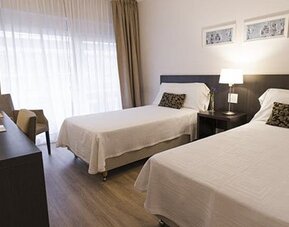 Twin-bed guest room with a desk, television, armchair, bedside table, and large curtained window at El Conquistador Hotel.