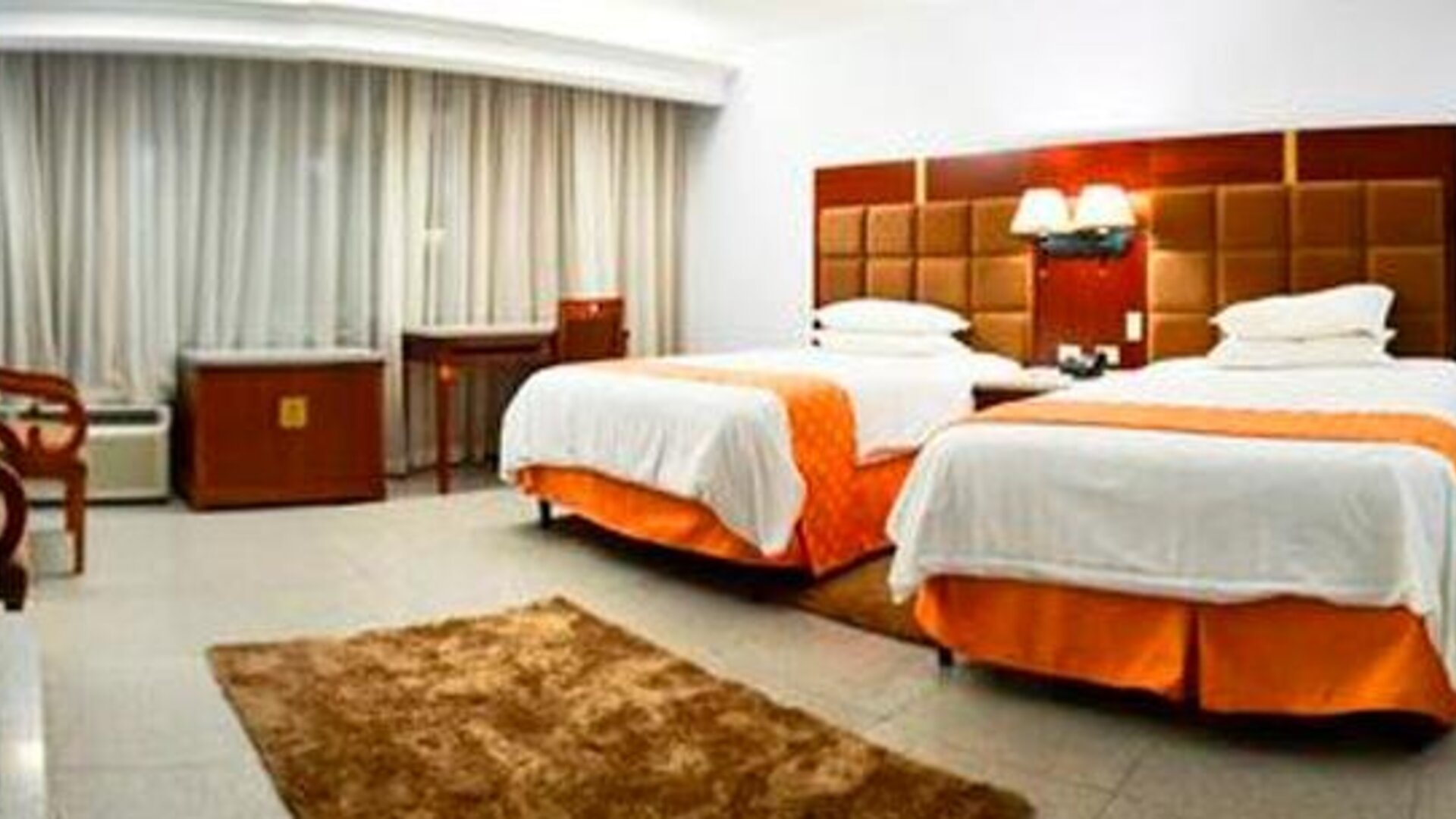 Guest room with two beds, orange runners, bedside lamps, desk, chair, and large curtained window at Hotel Grand International.