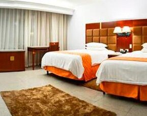 Guest room with two beds, orange runners, bedside lamps, desk, chair, and large curtained window at Hotel Grand International.