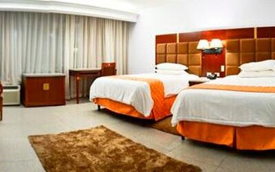 Guest room with two beds, orange runners, bedside lamps, desk, chair, and large curtained window at Hotel Grand International.