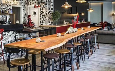 Hotel lounge and bar with a long wooden communal table, stools, sofas, pendant lights, and patterned walls at Hotel ibis Styles Barcelona City Bogatell.