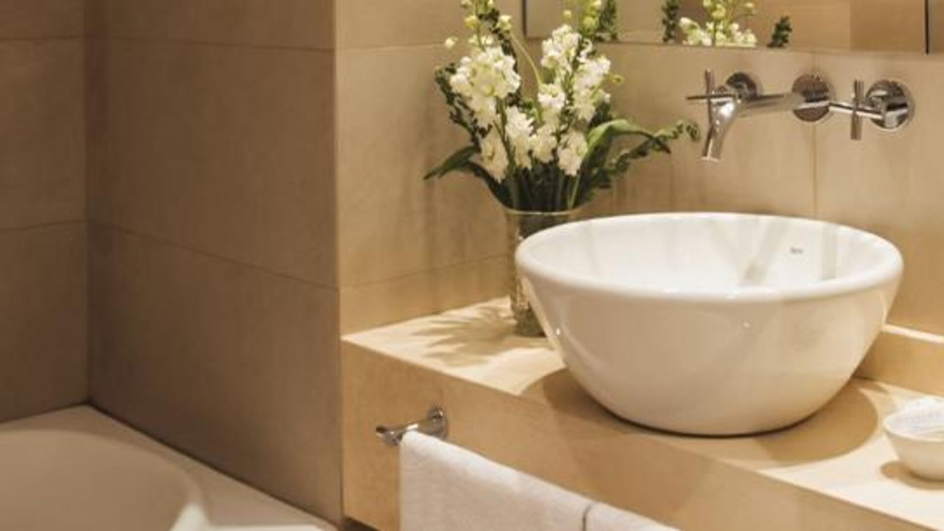 Bathroom with a white vessel sink, bathtub, wall-mounted faucet, toiletries, and floral arrangement at Dazzler by Wyndham Buenos Aires Recoleta.